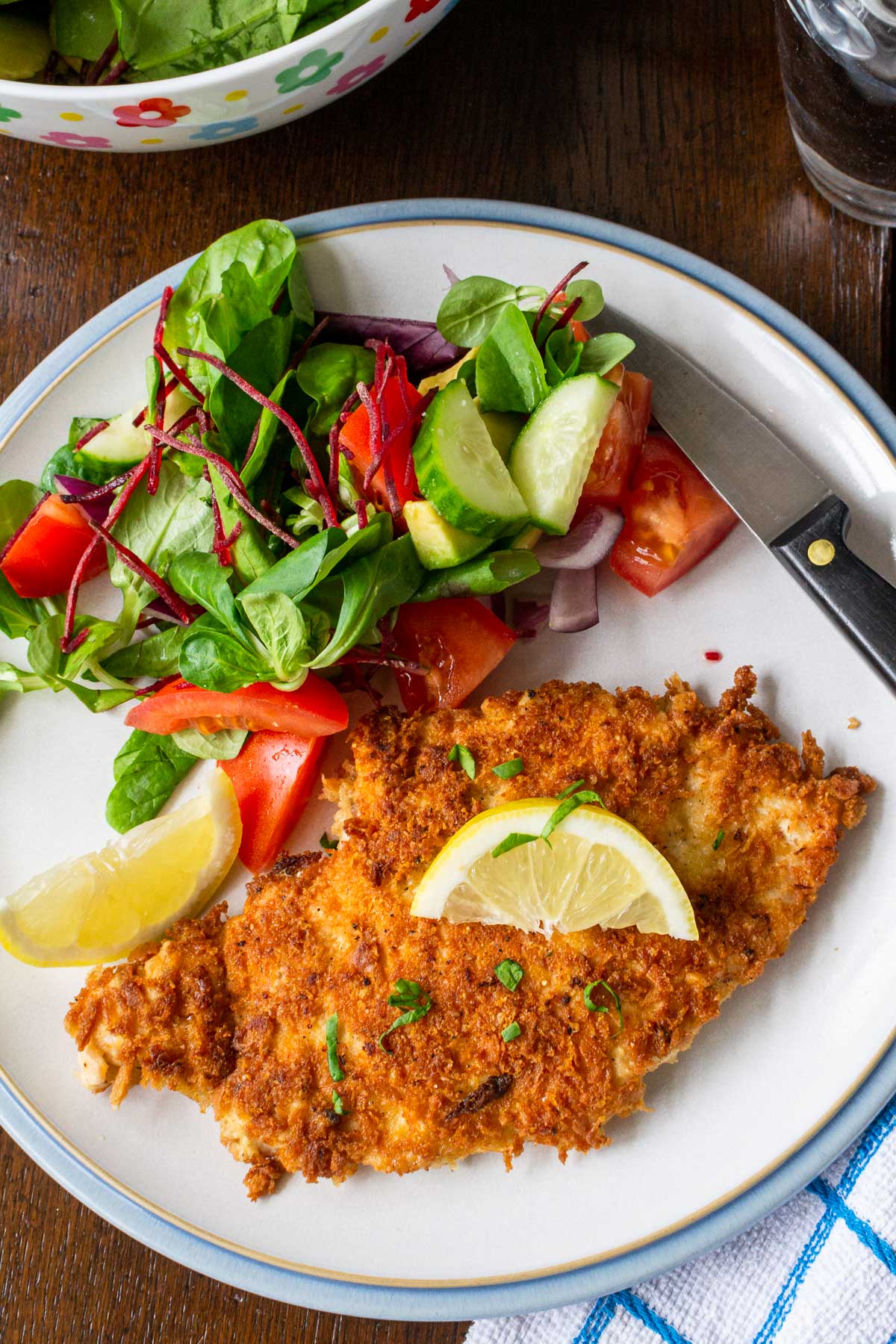 A crispy looking panko breadcrumbs crusted chicken breast on a plate with a blue rim from above with a simple side salad on the plate and with blue and white checked tea towel below on a wooden table.