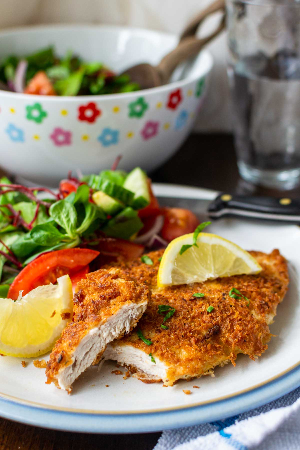 A piece of panko chicken with the end sliced off to reveal inside on a white plate with blue rim, with brightly colored salad bowl and water glass in background.