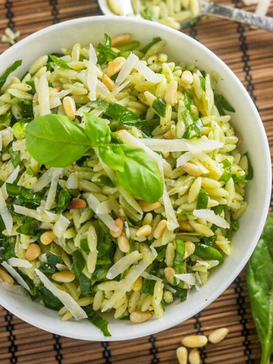 A closeup of an overhead view of a white bowl of pesto salad with orzo with grated parmesan and basil on top, all on a brown wicker background with a spoon in the background at the top.
