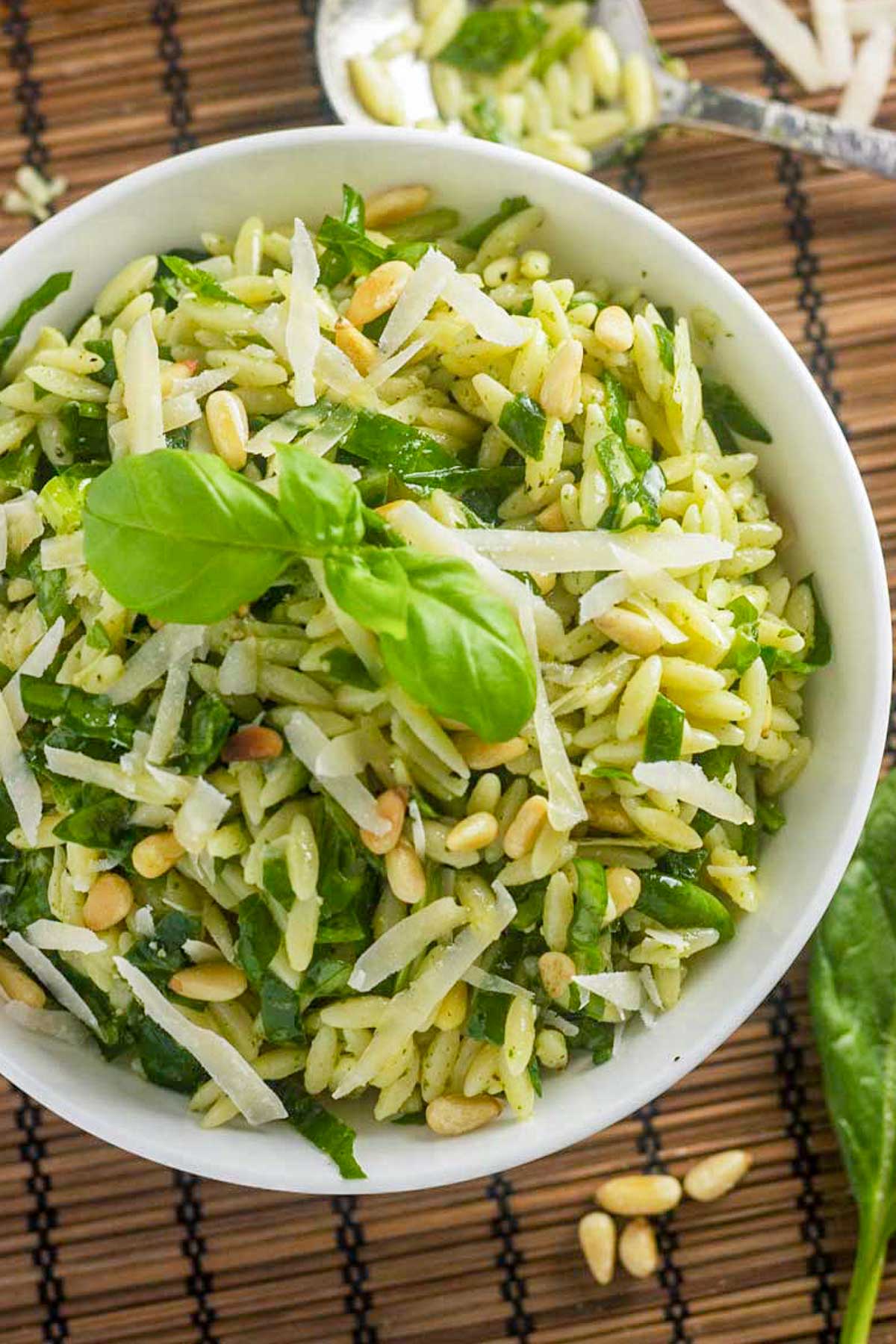 A closeup of an overhead view of a white bowl of pesto salad with orzo with grated parmesan and basil on top, all on a brown wicker background with a spoon in the background at the top.