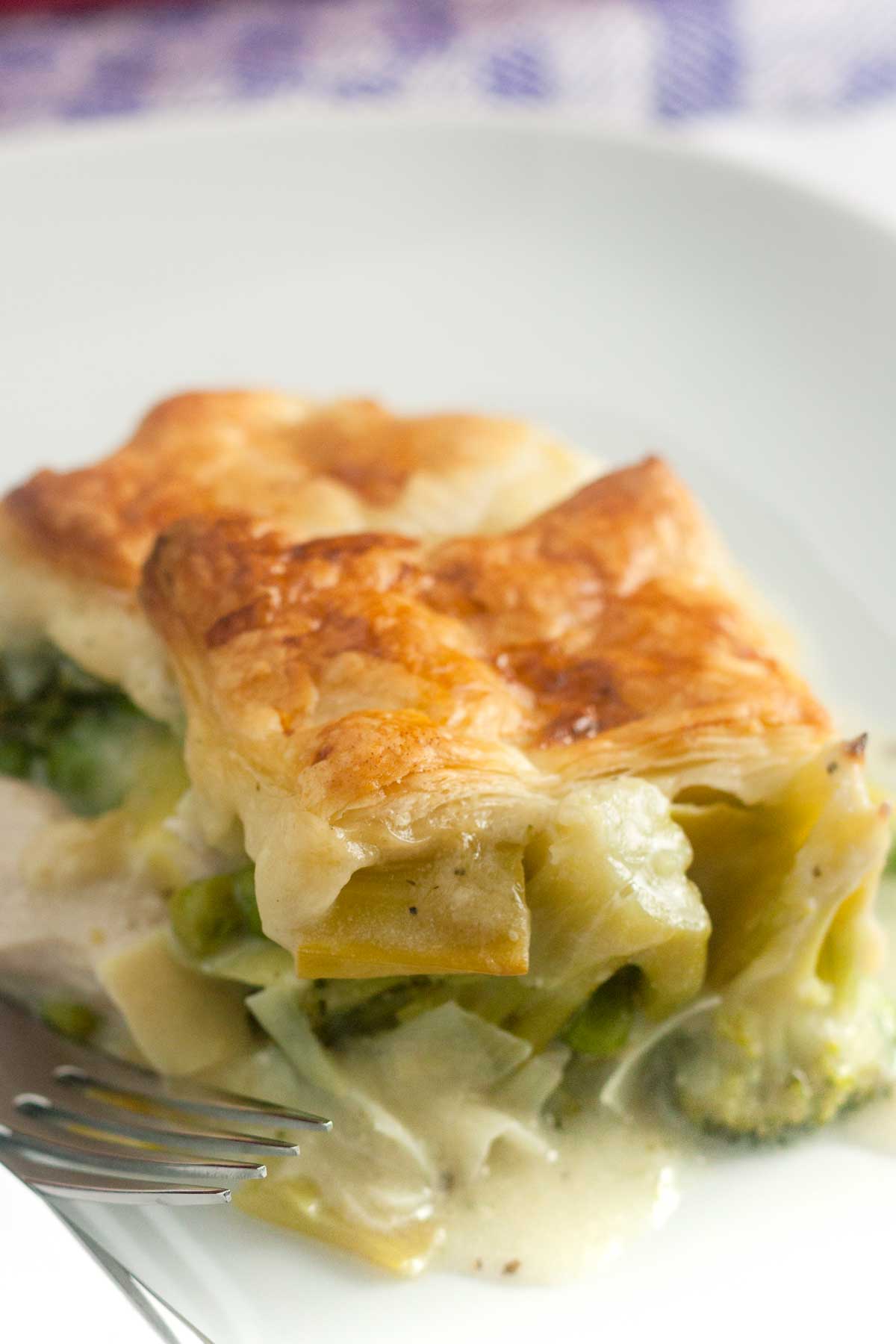 A piece of chicken and leek pie in a creamy sauce with a square of puff pastry on top, on a white plate with a fork.