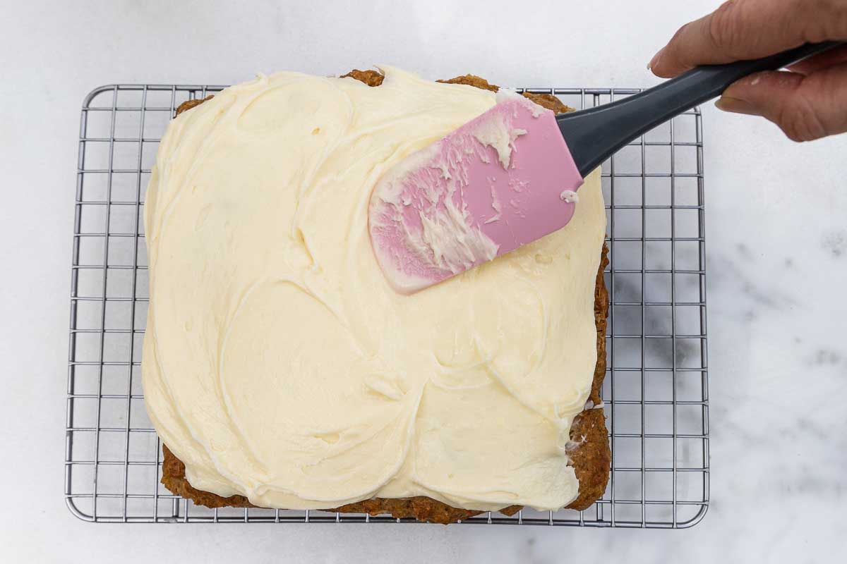 Overhead view of using pink spatula to spread cream cheese frosting onto pineapple banana cake on a cooling rack and marble background.