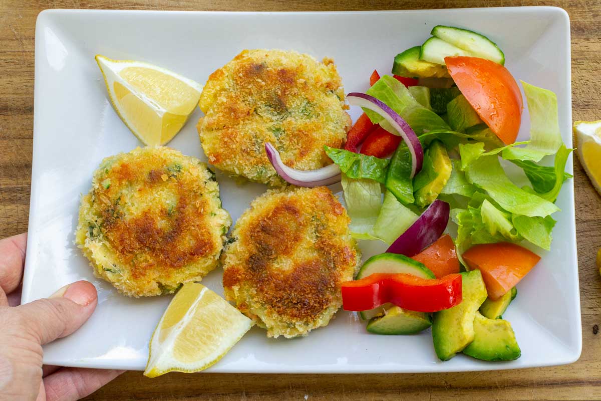 Someone holding a rectangular white plate with 3 salmon fishcakes, lemon segments and a simple salad on it.
