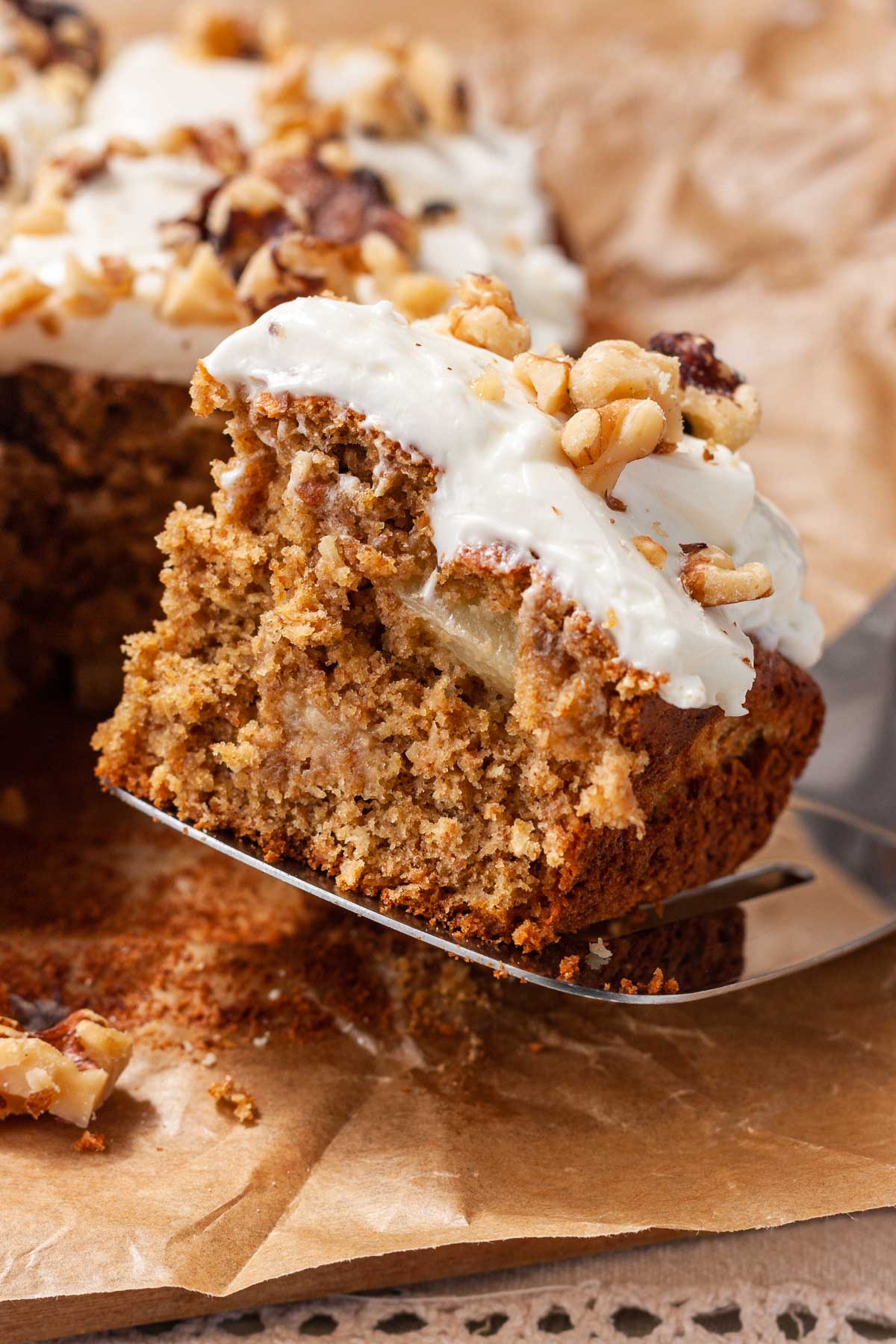 Closeup of a cake slice with a square piece of pineapple banana cake with cream cheese frosting and walnuts on top, on top of brown paper with the rest of the cake in the background.