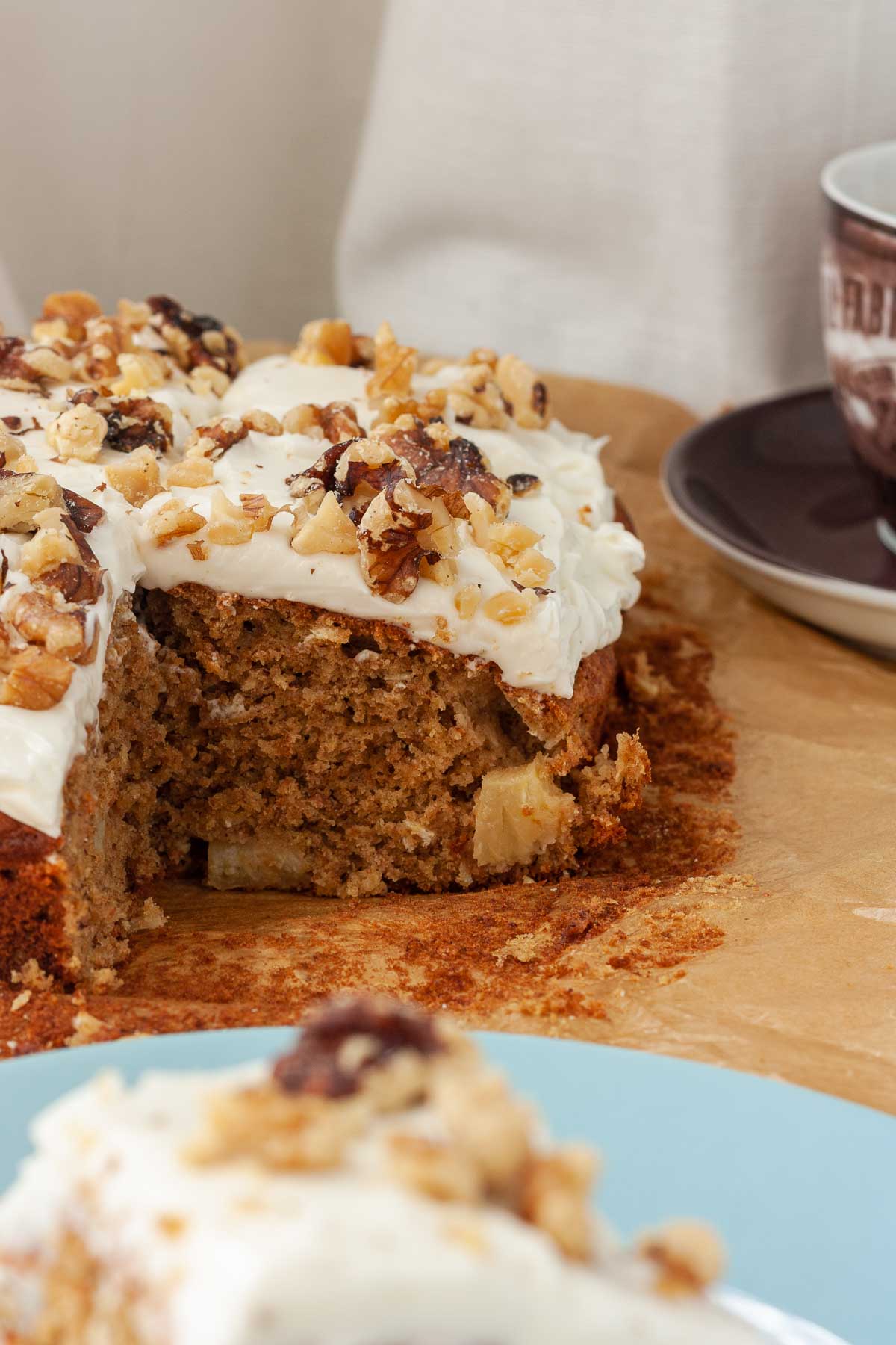 Closeup of the inside of a corner of cut pineapple banana cake with cream cheese frosting and walnuts on top, with another piece of cake in the foreground and a brown cup and saucer top right.