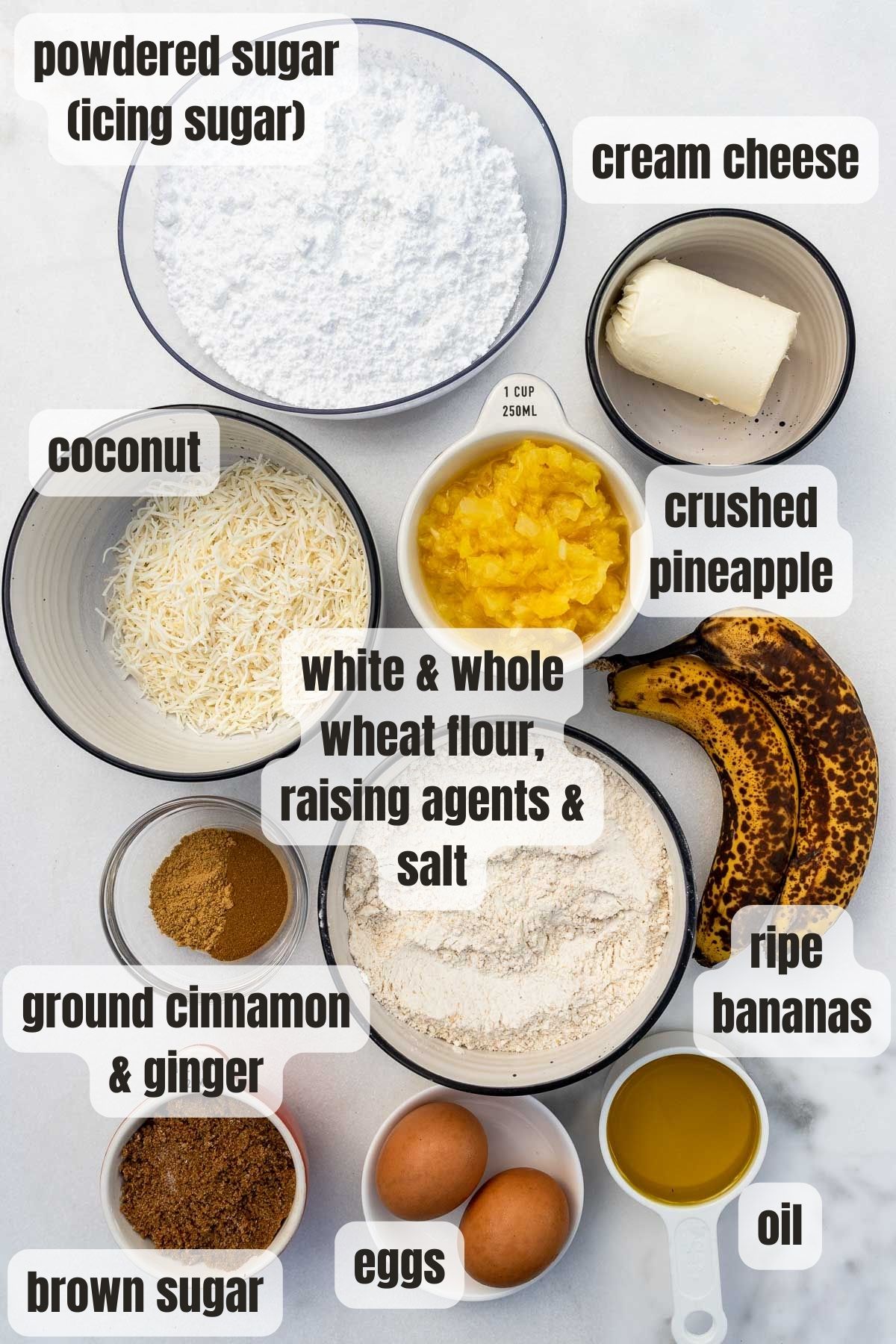 Overhead view of all the ingredients needed for a pineapple banana cake including white and whole wheat flour, raising agents and salt, brown sugar, eggs, oil, ripe bananas, ground cinnamon and ginger, coconut, crushed pineapple, cream cheese and powdered sugar.