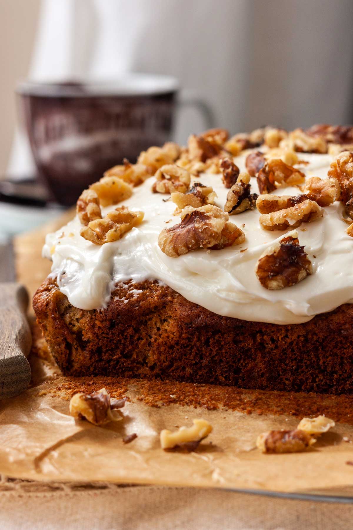 Closeup of the front part of a whole pineapple banana coconut cake with thick cream cheese frosting and walnuts on top on brown paper with walnuts in front and a brown cup and saucer behind.