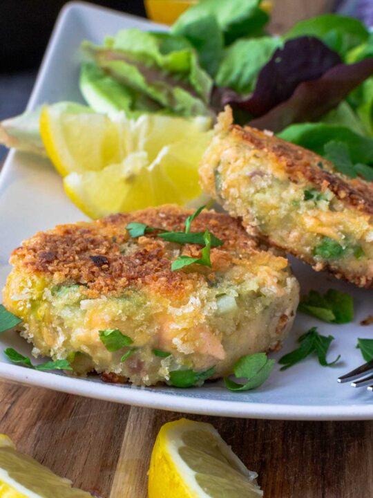 Closeup of 2 crispy salmon fish cakes on a rectangular white plate with green salad in the background and lemon segments on a wooden board in the foreground.
