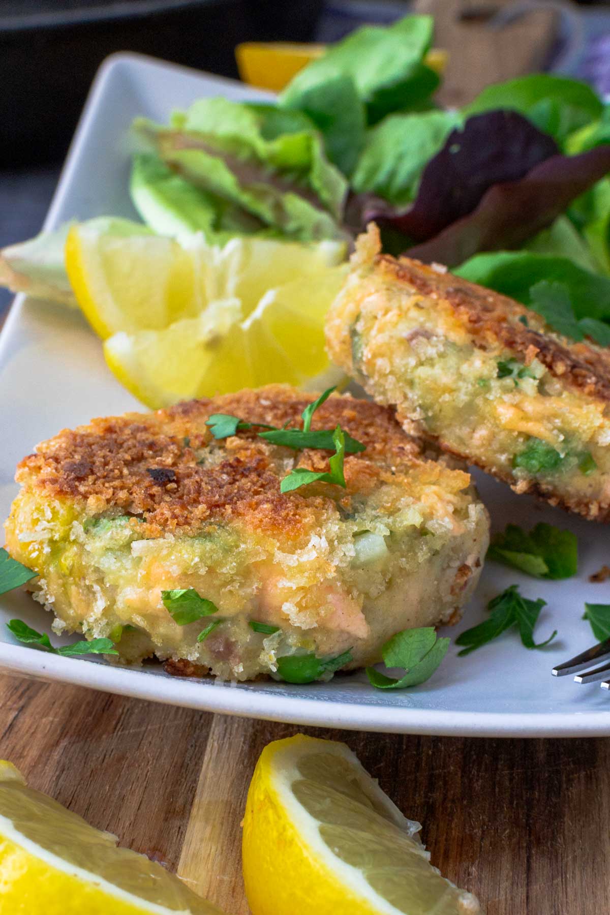 Closeup of 2 crispy salmon fish cakes on a rectangular white plate with green salad in the background and lemon segments on a wooden board in the foreground.