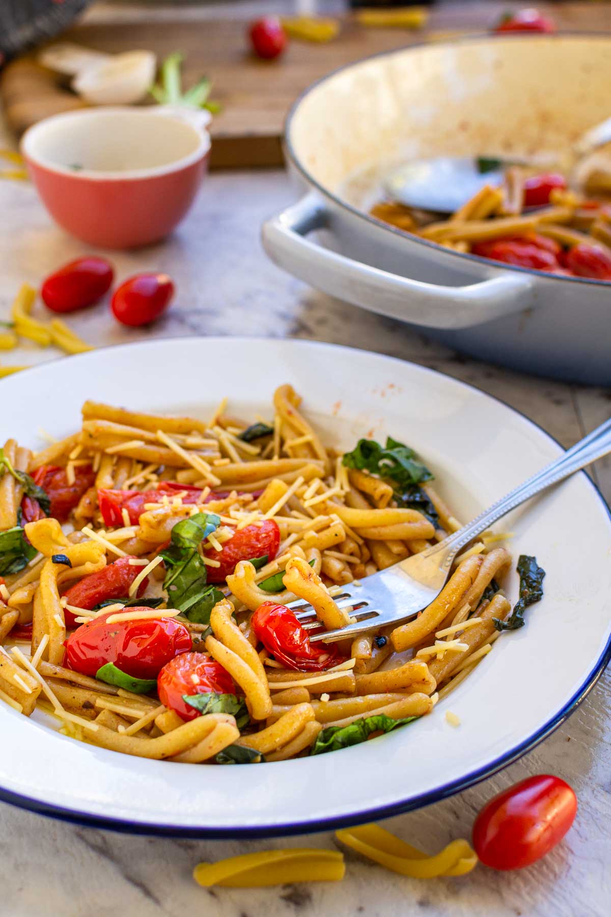 A fork in a white and blue plate of casarecce pasta with tomatoes and basil and parmesan with the blue cast iron pan, chopping board and ingredients in the background.