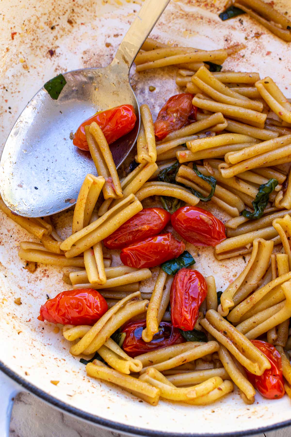 Closeup of overhead of casarecce pasta and cherry tomatoes and basil in a blue cast iron pan on a marble background and with a serving spoon in it.
