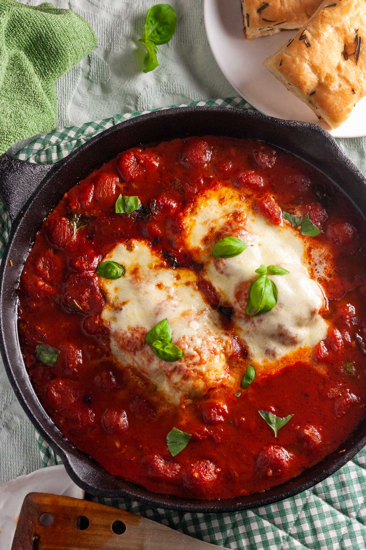 Overhead view of chicken pizzaiola or chicken breasts in a cherry tomato and basil sauce with bubbling mozzarella on top on a green and white checked cloth with focaccia and a wooden spoon at the side.