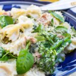 Closeup of a dish of farfalle pasta with prosciutto, broccoli and ricotta with cutlery in a blue and white dish on a marble background and with half a lemon in the background.