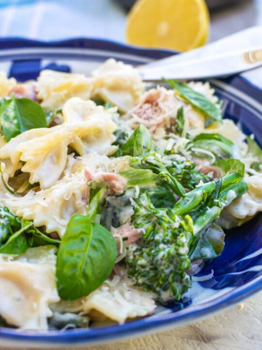 Closeup of a dish of farfalle pasta with prosciutto, broccoli and ricotta with cutlery in a blue and white dish on a marble background and with half a lemon in the background.