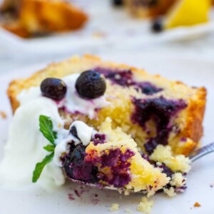 A slice of lemon curd blueberry cake on a white plate with Greek yogurt and blueberries on top and with a piece of the cake on a fork.