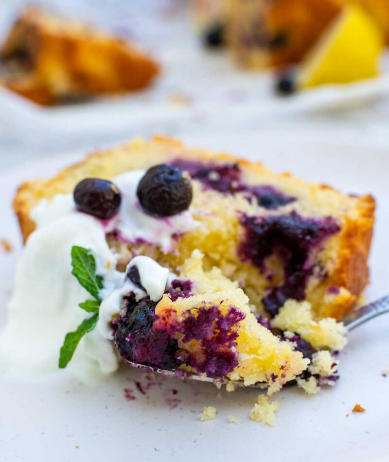 A slice of lemon curd blueberry cake on a white plate with Greek yogurt and blueberries on top and with a piece of the cake on a fork.