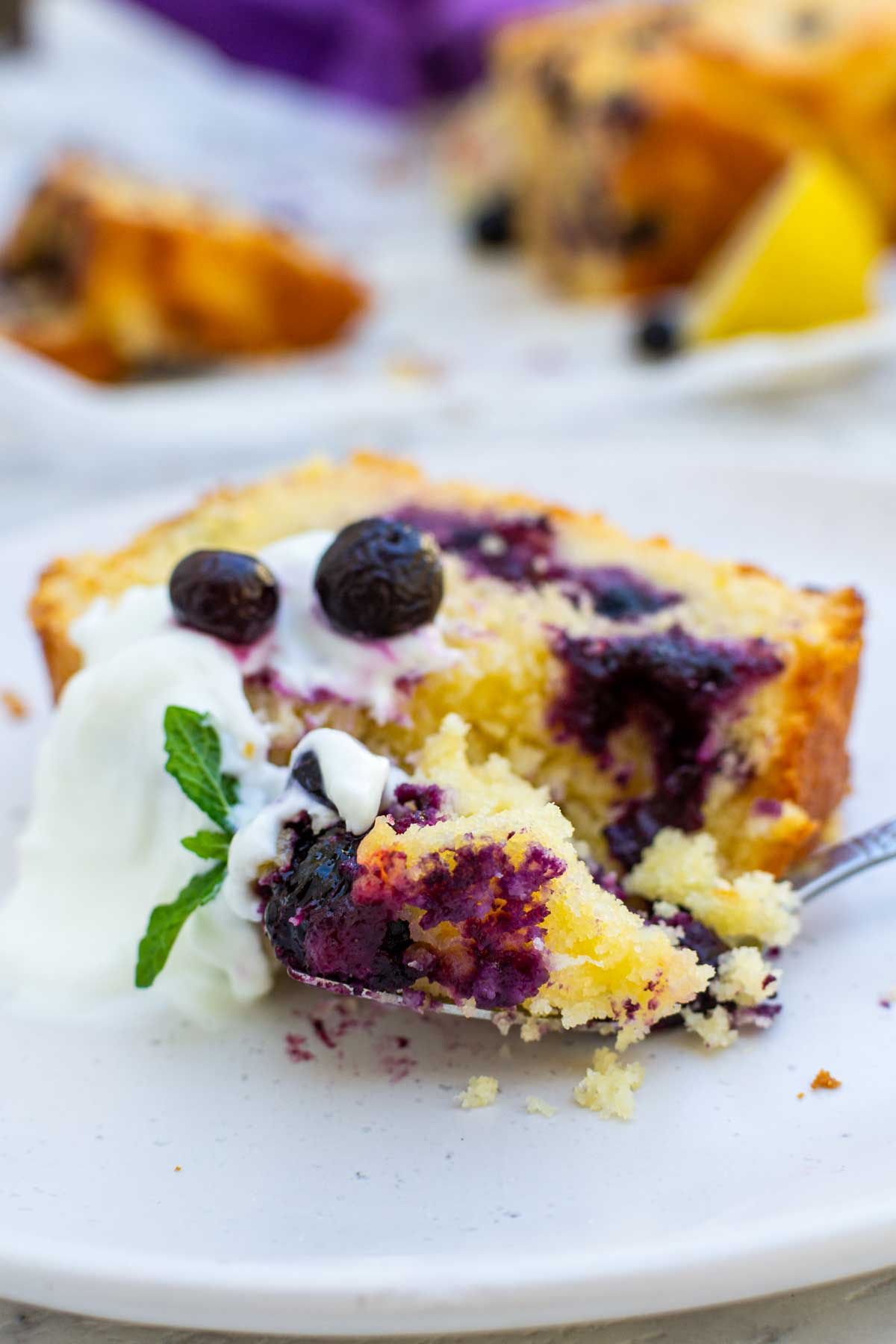 A slice of lemon curd blueberry cake on a white plate with Greek yogurt and blueberries on top and with a piece of the cake on a fork.
