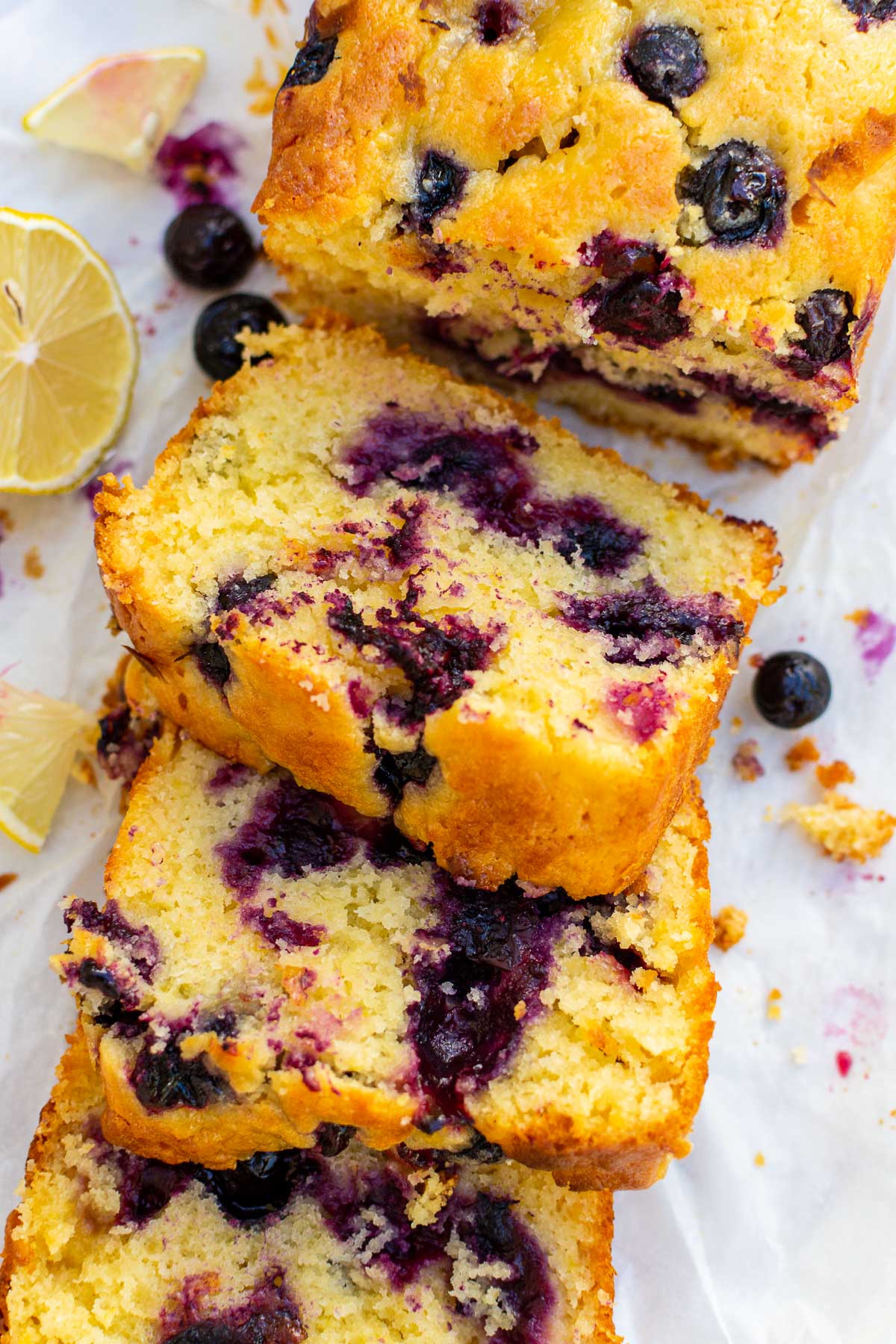 Closeup overhead shot of slices of lemon curd blueberry cake on baking paper with a few extra blueberries and lemon quarters at the side.