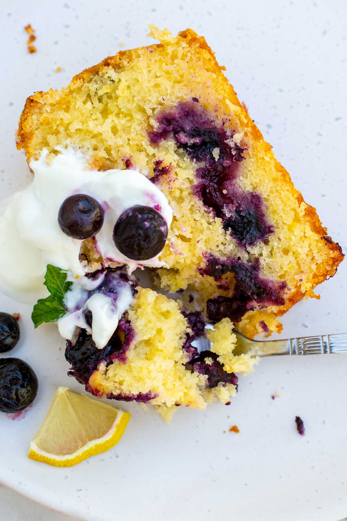 Overhead view of a slice of lemon curd cake with blueberries with Greek yogurt on top on a white plate with a fork.