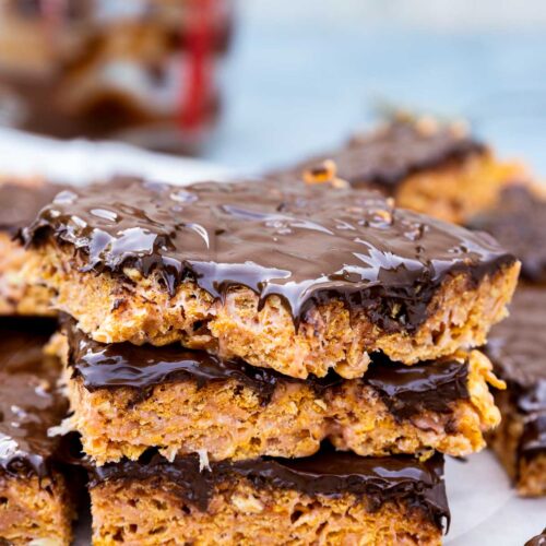 Closeup of 3 peanut butter cornflake bars on top of each other with a bite out of the top one, all on baking paper with more bars at the side and a few cornflakes and with the empty melted chocolate jug in the background.