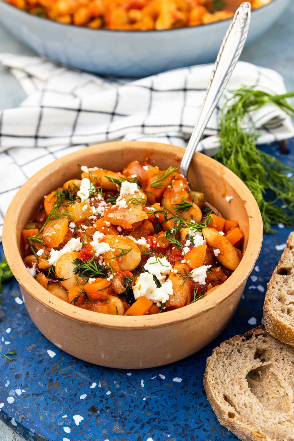 A ceramic dish of Greek giant beans with fresh herbs and feta crumbled on top, on a blue marble plate stand with slices of bread, fresh dill, a white and blue checked tea towel and a bigger pan of the beans around it.