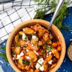 An overhead view of a ceramic dish of Greek giant beans with fresh herbs and feta crumbled on top, on a blue marble plate stand with slices of bread, fresh dill, a white and blue checked tea towel and a bigger pan of the beans around it.
