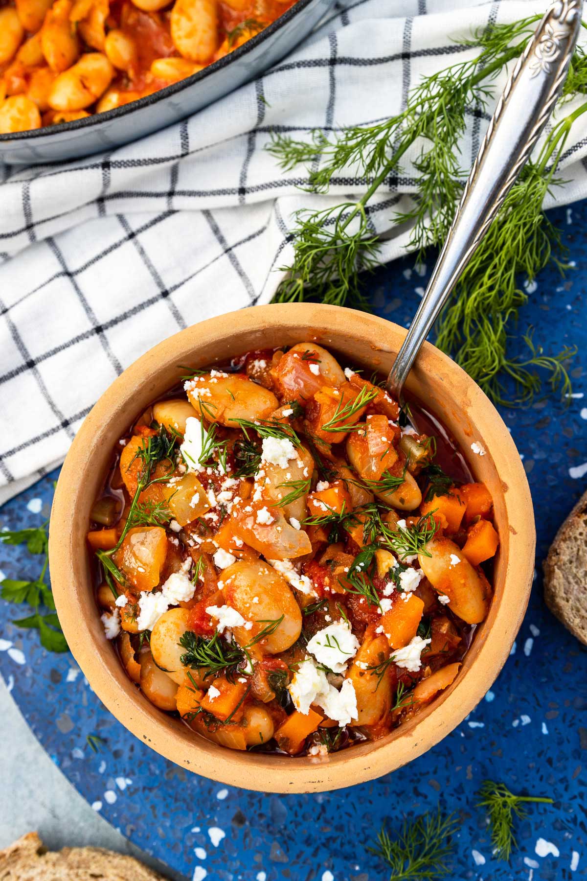 An overhead view of a ceramic dish of Greek giant beans with fresh herbs and feta crumbled on top, on a blue marble plate stand with slices of bread, fresh dill, a white and blue checked tea towel and a bigger pan of the beans around it.