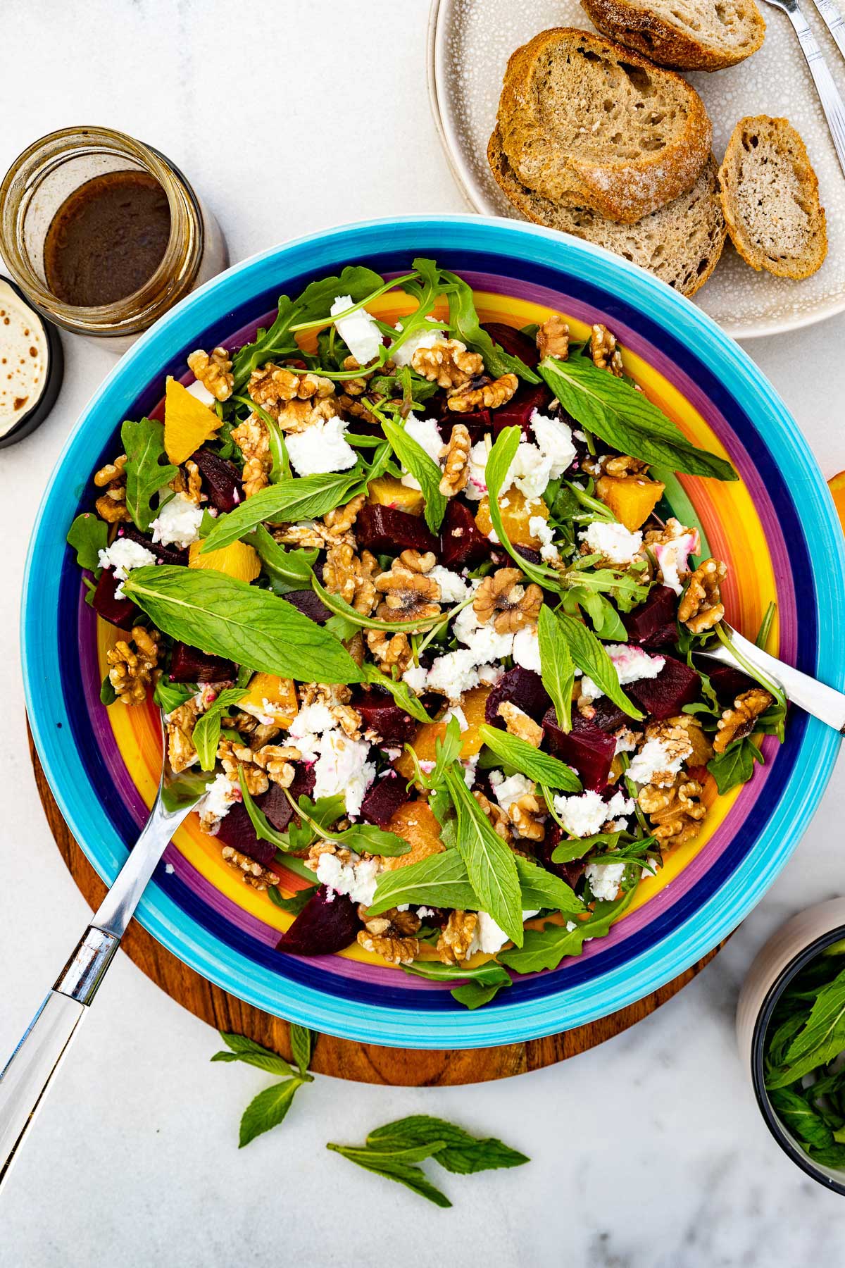 A colorful dish of beetroot and feta salad with servers in it on a wooden board and marble background with fresh mint scattered around and a jar of dressing and plate of bread at the top.