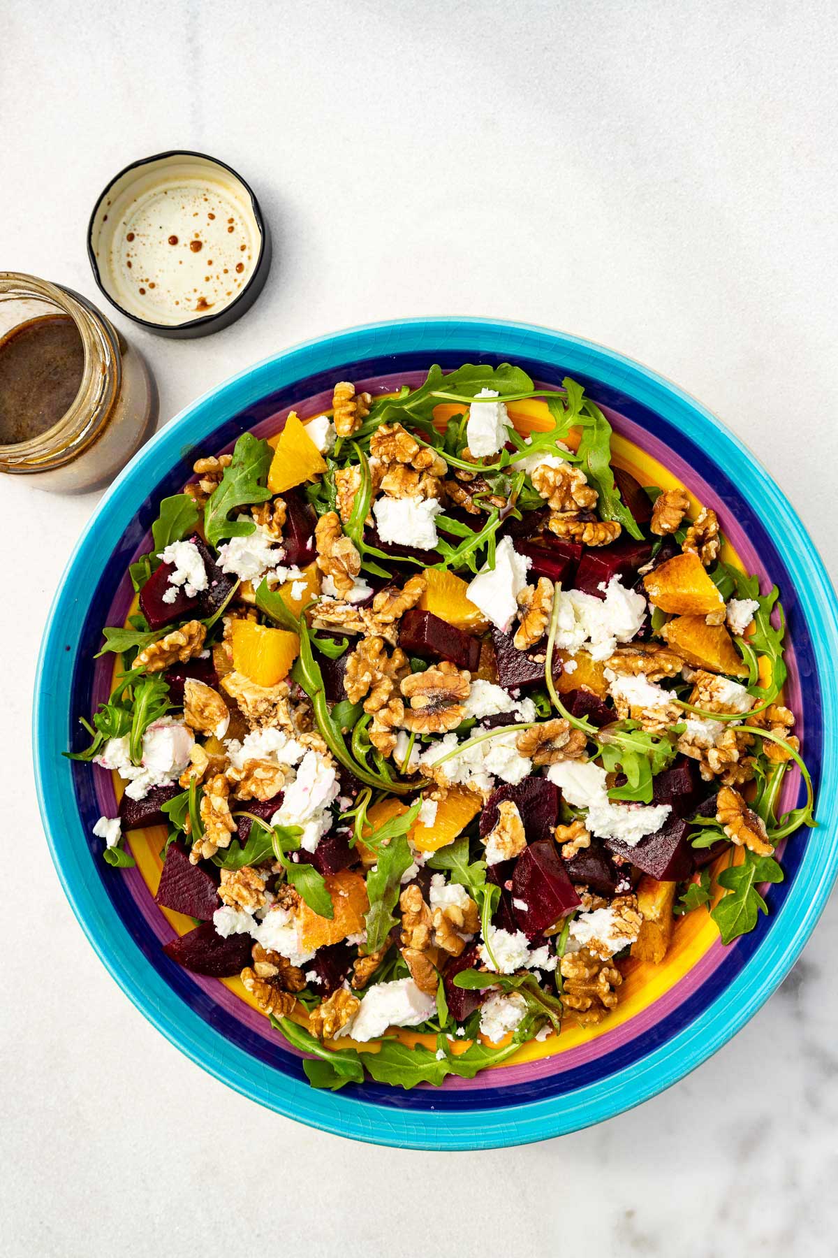 A beetroot and feta salad with walnuts and orange in a colorful salad bowl on a marble background with an opened jar of dressing top left.