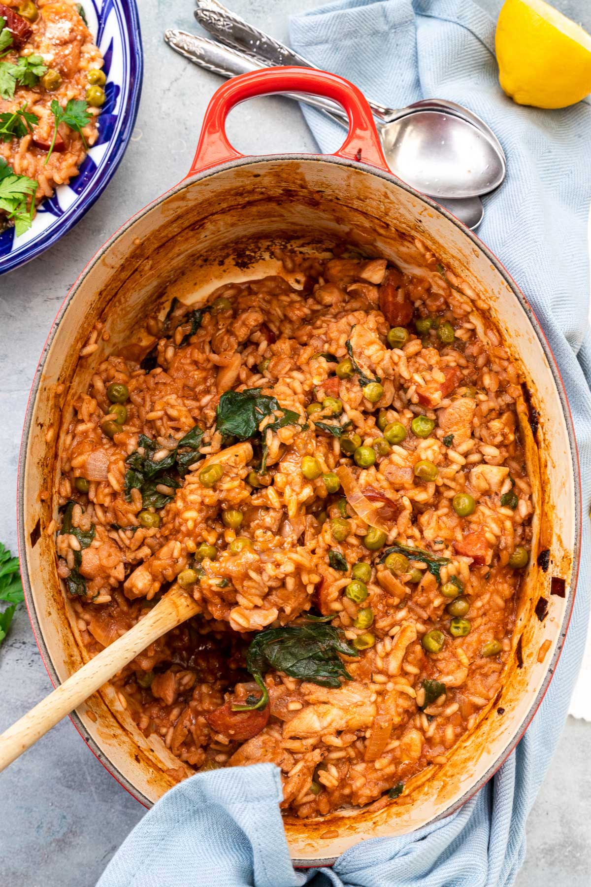 Overhead of a red cast iron pan with chicken and chorizo risotto in it with a wooden spoon in it and a pale blue tea towel wrapped around it, all on a blue background.