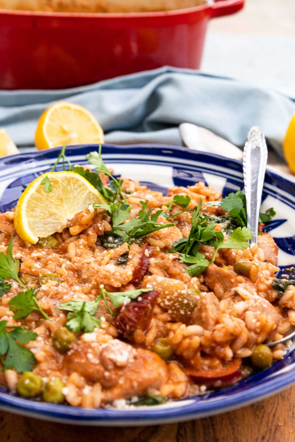 Closeup of a a blue patterned dish of chicken and chorizo risotto with a spoon in and lemon and herbs and parmesan on top, all on a wooden round board and cloth background with equipment and ingredients behind.