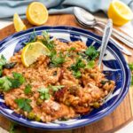 Closeup of a blue patterned dish of chicken and chorizo risotto with a spoon in and lemon and herbs and parmesan on top, all on a wooden round board and cloth background with equipment and ingredients behind.
