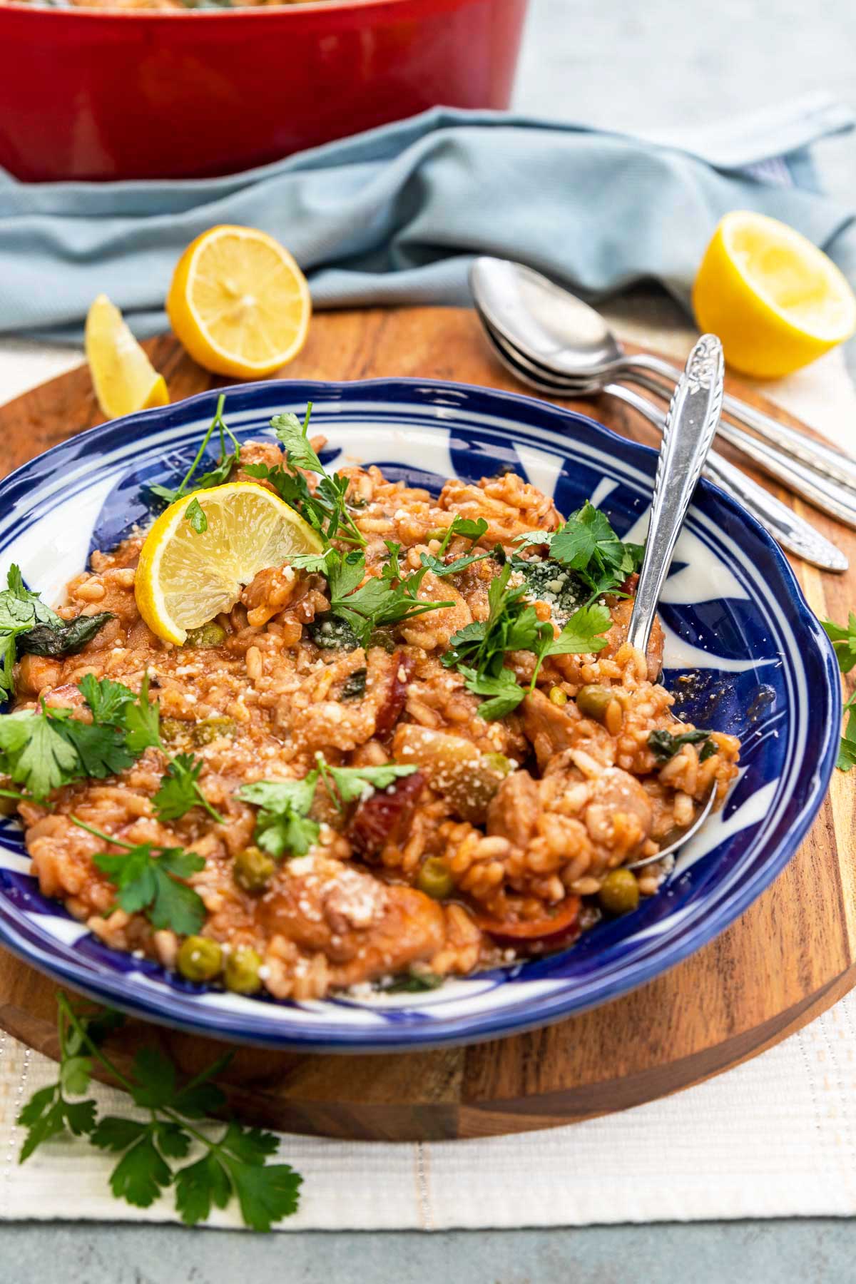 Closeup of a blue patterned dish of chicken and chorizo risotto with a spoon in and lemon and herbs and parmesan on top, all on a wooden round board and cloth background with equipment and ingredients behind.