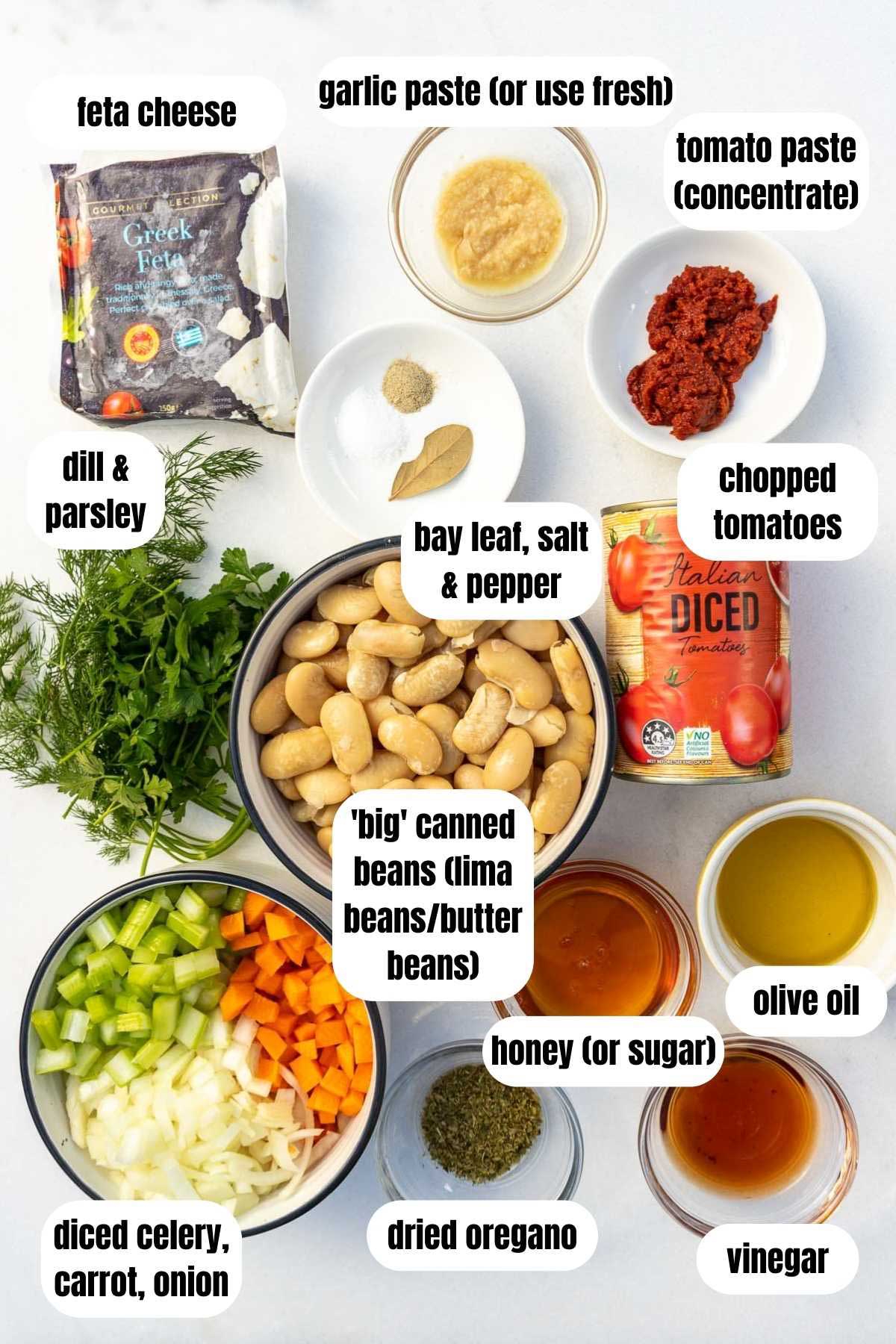 An overhead view of all the ingredients needed to make quick Greek giant beans including chopped onion, carrot and celery, dried oregano, garlic paste, vinegar, honey, olive oil, canned lima or butter beans, chopped tomatoes, tomato paste, salt, pepper and a bay leaf, feta cheese, and fresh dill and parsley.