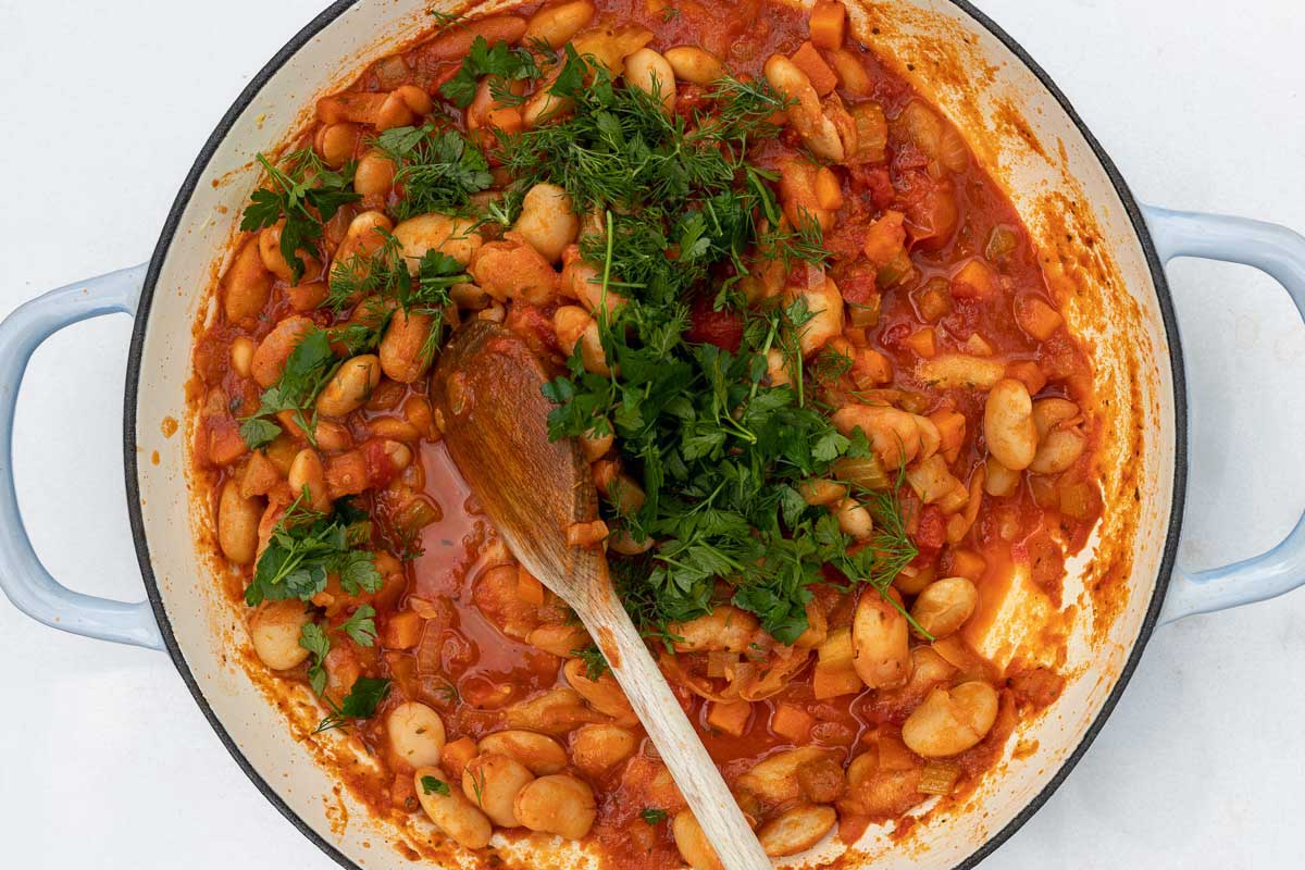 Overhead of a blue cast iron pan on a marble background with cooked Greek beans in tomato sauce and a wooden spoon and with fresh dill and parsley on top ready to be stirred in.