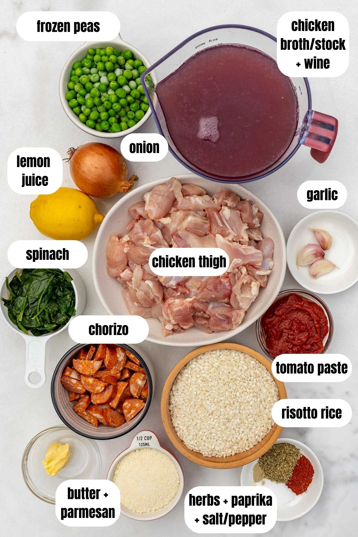 Overhead of all the ingredients for a chicken chorizo risotto including risotto rice, chicken broth, butter, parmesan cheese, herbs and spices, tomato paste, chicken thigh, spinach, garlic, onion, lemon juice and frozen peas.