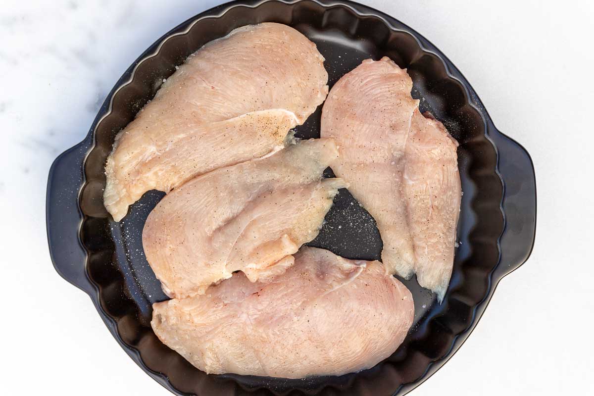 Overhead of thin raw chicken breasts with salt and pepper in a round black baking dish on a marble background.