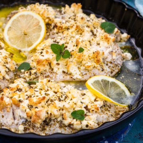 Most of a round black baking dish with thin chicken breasts baked with lemon juice, olive oil, oregano and lots of crumbled feta, all on a weathered blue background with lemon segments, a fresh oregano sprig and a blue tea towel around the dish.