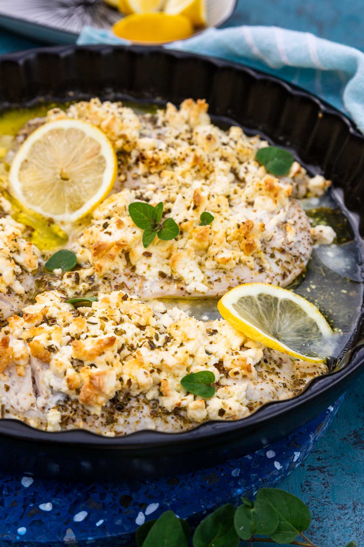 Most of a round black baking dish with thin chicken breasts baked with lemon juice, olive oil, oregano and lots of crumbled feta, all on a weathered blue background with lemon segments, a fresh oregano sprig and a blue tea towel around the dish.