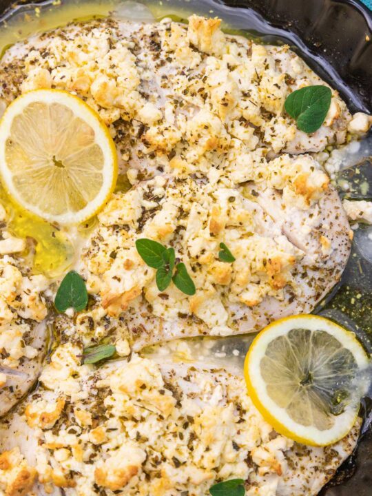 A closeup of part of a round black baking dish with thin chicken breasts baked with lemon juice, olive oil, oregano and lots of crumbled feta, with lemon slices and fresh oregano scattered on top.