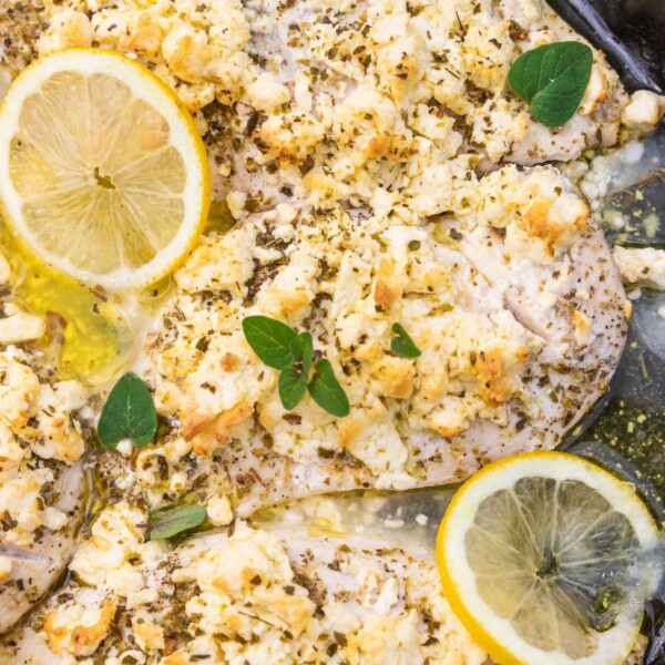A closeup of part of a round black baking dish with thin chicken breasts baked with lemon juice, olive oil, oregano and lots of crumbled feta, with lemon slices and fresh oregano scattered on top.