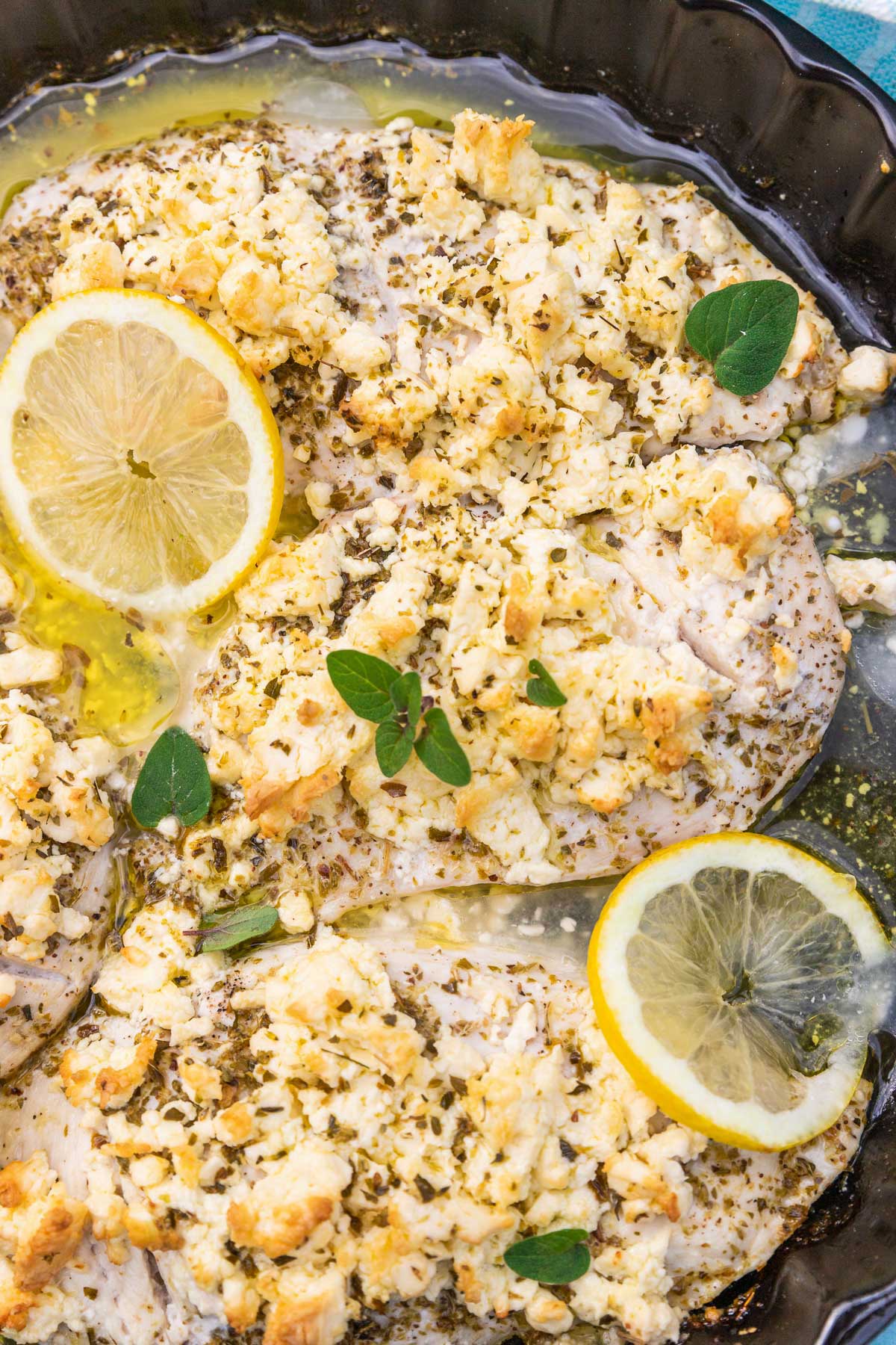 A closeup of part of a round black baking dish with thin chicken breasts baked with lemon juice, olive oil, oregano and lots of crumbled feta, with lemon slices and fresh oregano scattered on top.