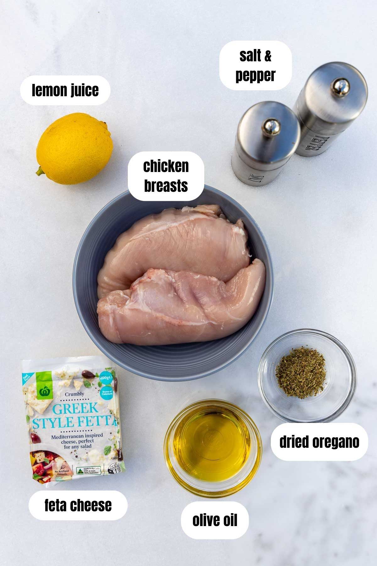 All the ingredients to make baked chicken breasts with lemon and feta labelled on a marble background, including chicken breasts, Greek feta in a package, olive oil, dried oregano, a lemon and salt and pepper.