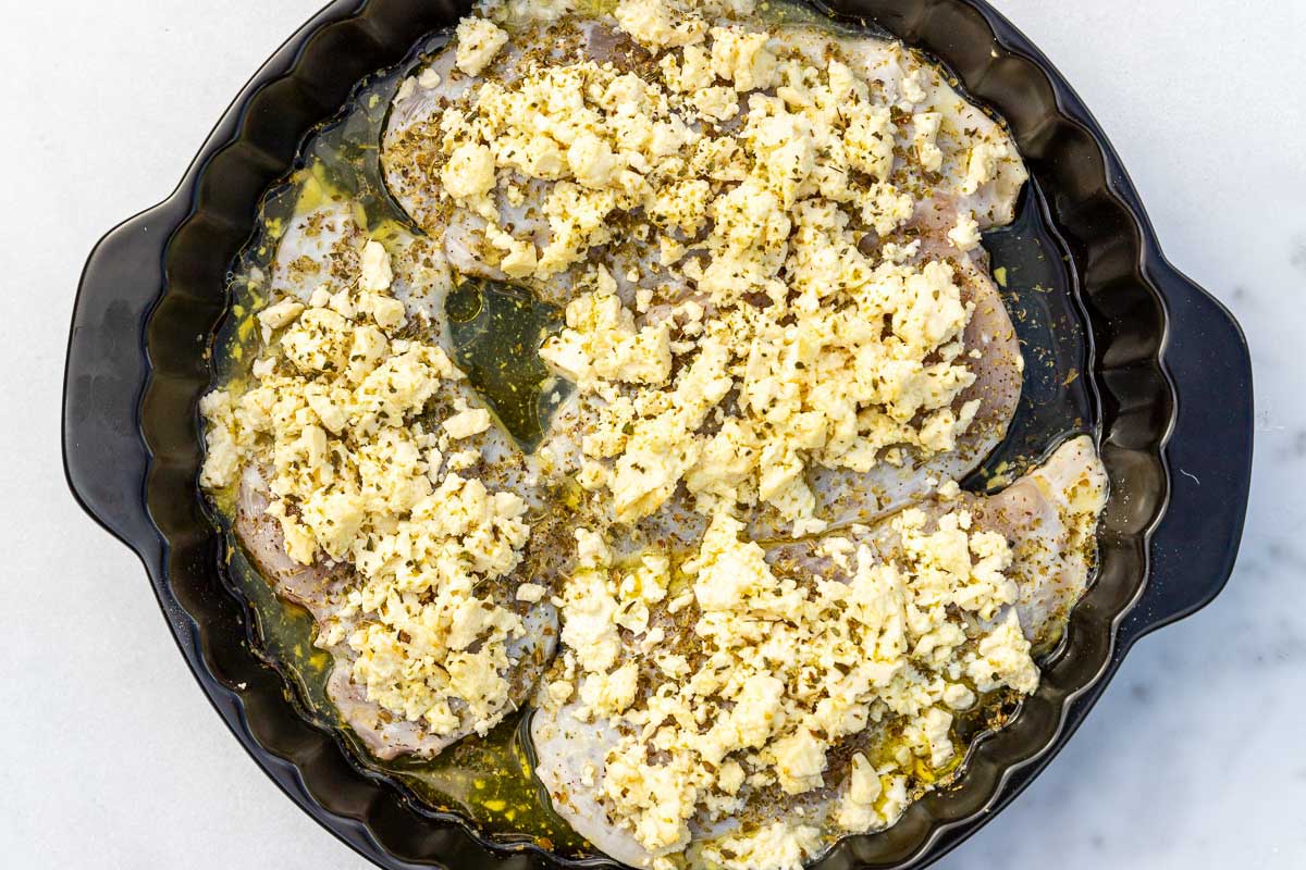 Raw thin chicken breasts laid in a round black baking dish with olive oil, lemon juice, oregano and lots of feta on top, all on a marble background.