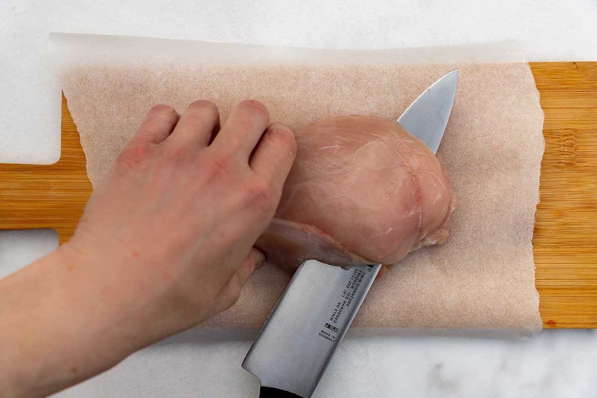 Overhead of someone's hand cutting a chicken breast in half across the width with a very big sharp knife on baking paper and a wooden chopping board.