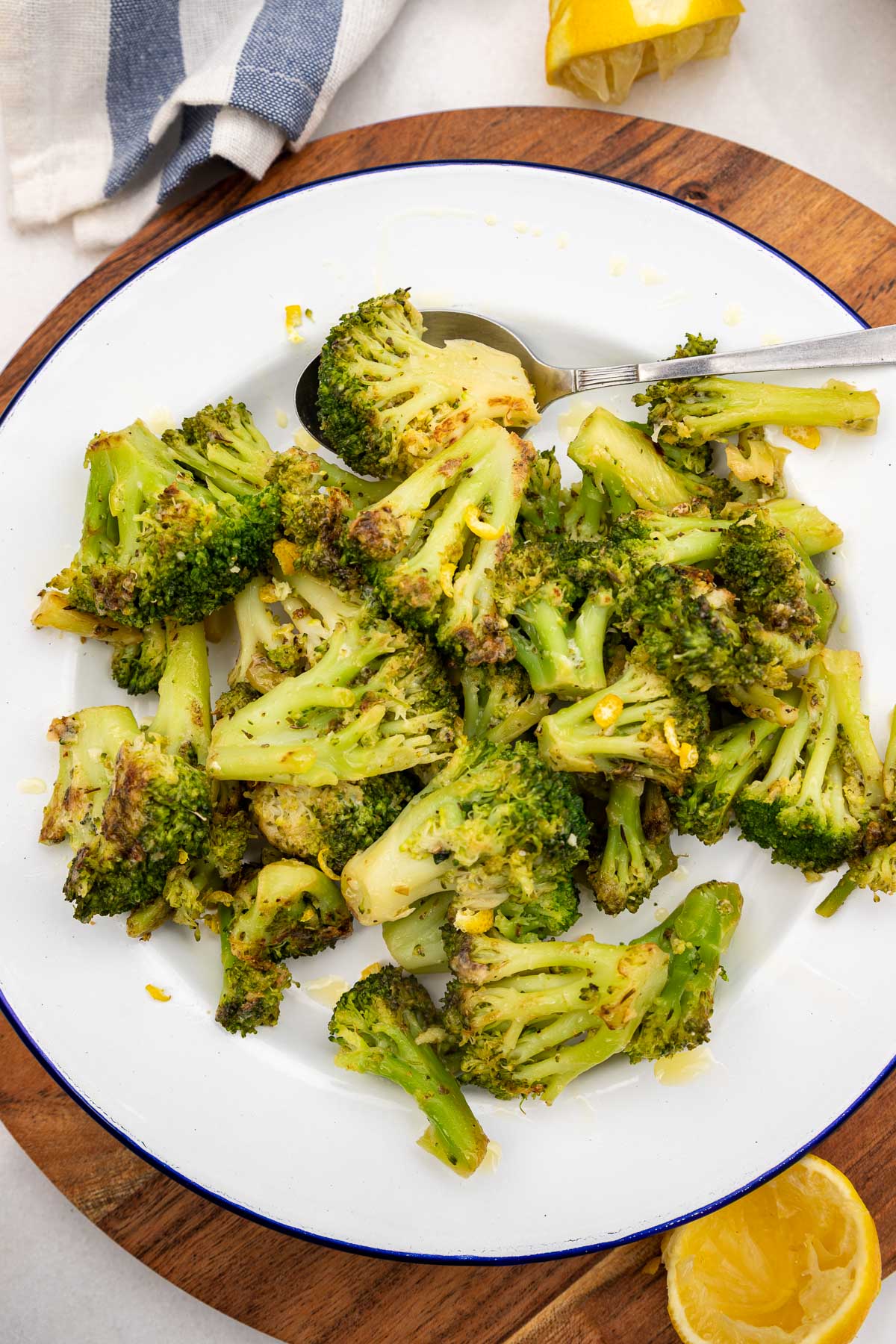 Overhead view of a plate of sauteed broccoli with lemon and parmesan on a white plate with blue rim on a round wooden board and with 2 squeezed lemon halves at the side and a blue and white tea towel at the top.