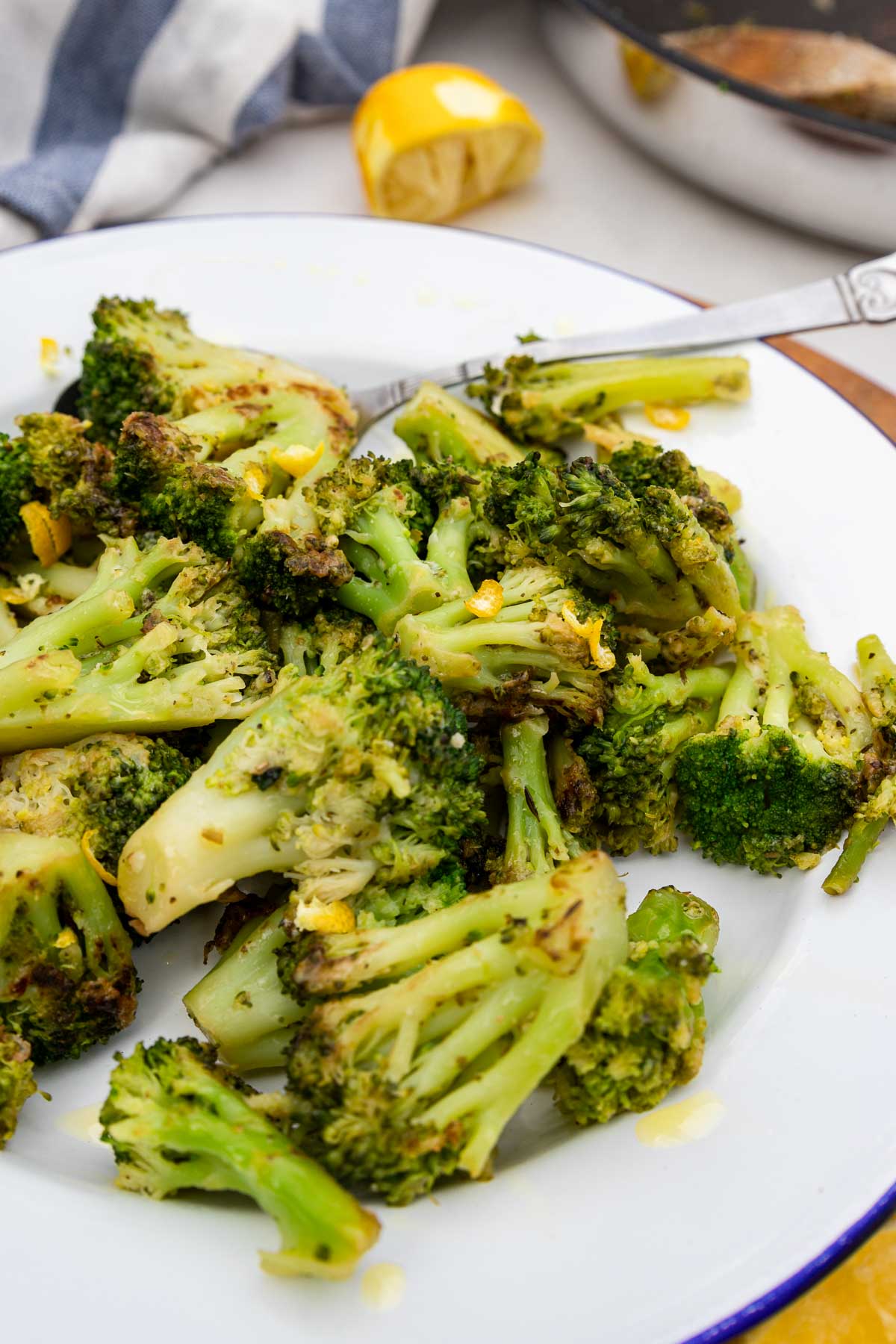 Overhead view of a closeup of a plate of sauteed broccoli with lemon and parmesan on a white plate with blue rim on a round wooden board and with a lemon half at the top and a blue and white tea towel.