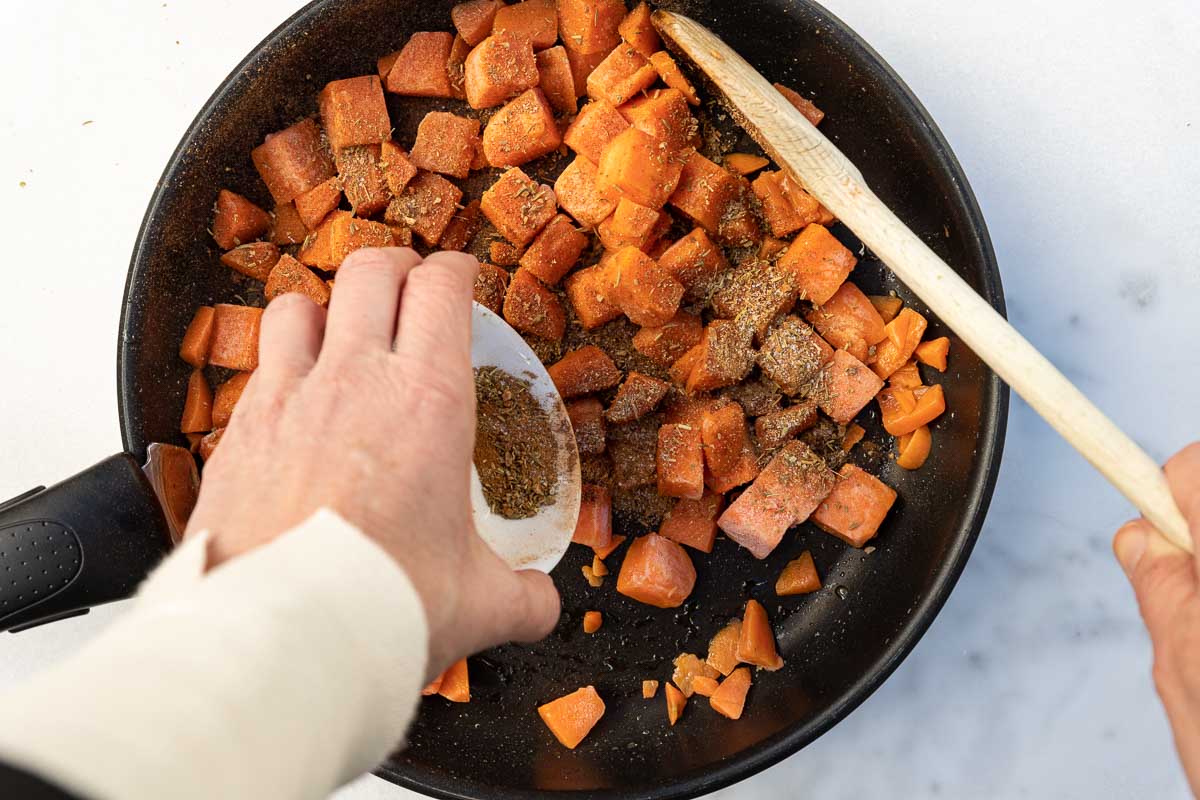 Someone's hand adding a small white pot of mixed herbs and spices to a frying pan of cubed butternut squash with a wooden spoon and on a marble background.