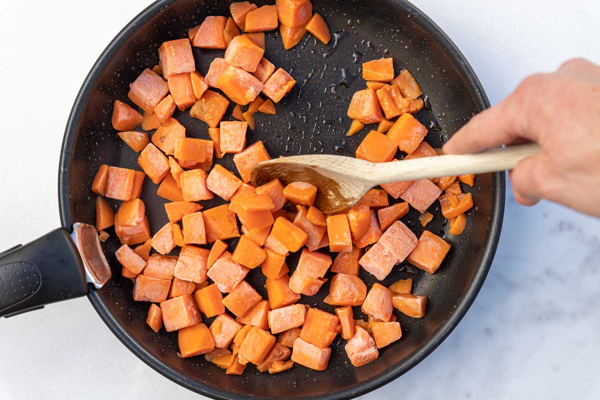 Overhead of someone's hand stirring a frying pan of cubed frozen butternut squash with a wooden spoon, all on a marble background.