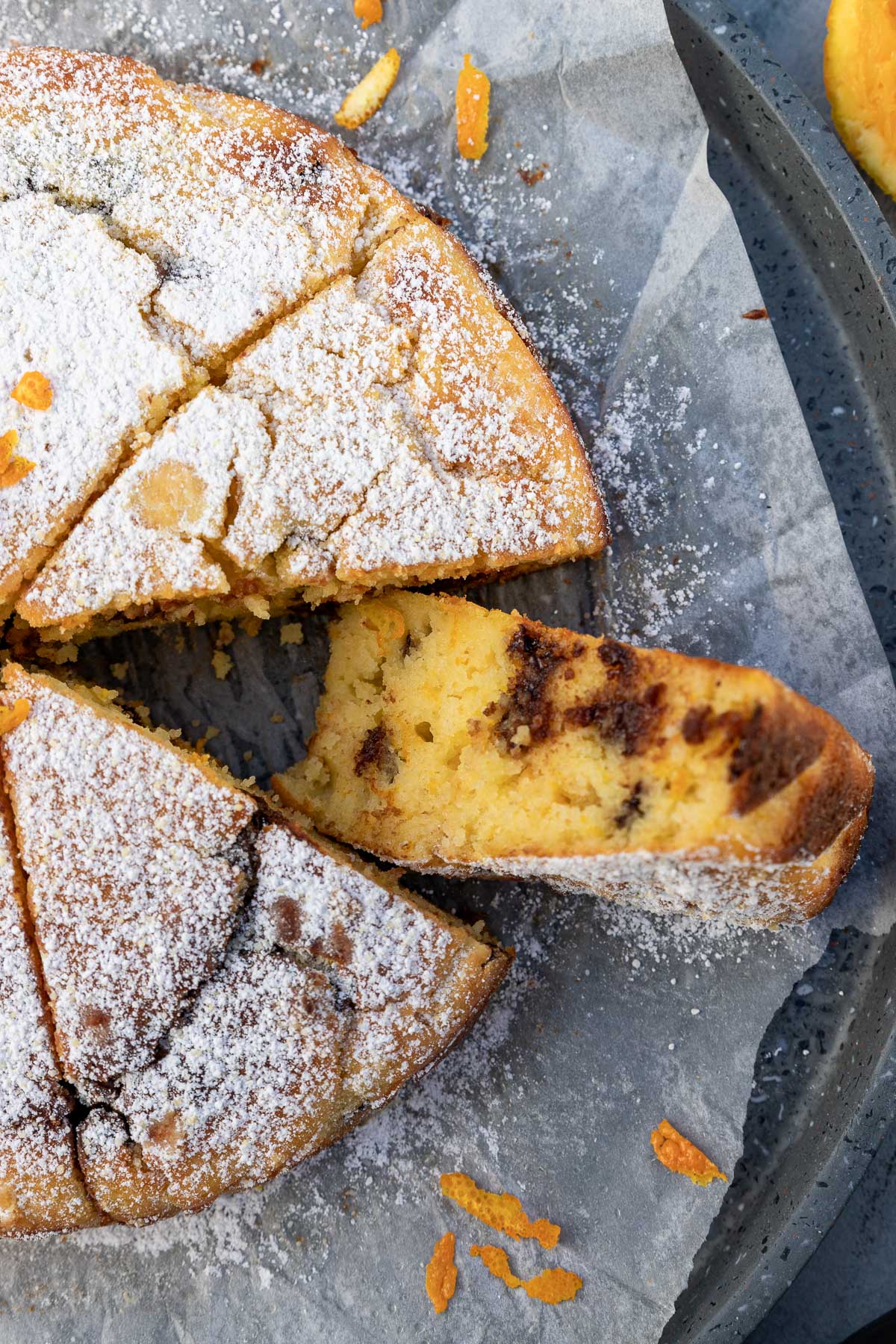 Overhead and closeup of part of a whole orange ricotta cake with chocolate chips with one piece on its side, all on baking paper and a round grey terrazo tray.